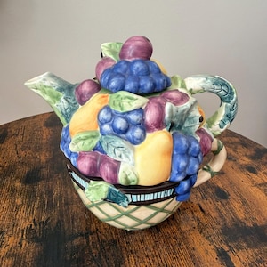 May include: A ceramic teapot shaped like a basket overflowing with sculpted fruit. The base features a green lattice pattern, while the top is adorned with clusters of blue grapes, purple plums, and yellow pears, complete with green leaves.