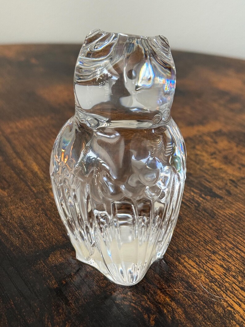 May include: A clear glass figurine shaped like an owl. The object features a smooth, rounded head with subtle indentations for eyes and a textured body with vertical ridges that mimic feathers, creating a reflective and translucent appearance.
