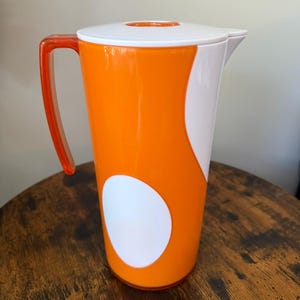 Puede incluir: Una jarra cilíndrica alta que presenta un cuerpo de color naranja vibrante con un gran acento ovalado blanco en la parte frontal. Incluye una tapa blanca, un asa de color naranja translúcido y un pico vertedor de material plástico sólido.