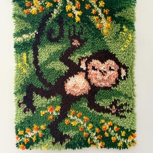 Könnte beinhalten: Handgefertigte, strukturierte Wandbehänge mit einem Affenmotiv. Der Affe ist braun und hellbraun, vor einem grünen Hintergrund mit einem Baum und bunten Blumen. Das Kunstwerk ist rechteckig und hat ein weiches, taktiles Aussehen.