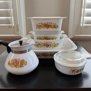 May include: A collection of white ceramic cookware with a colourful fruit and vegetable design. Includes various baking dishes, a teapot, and a covered bowl. The set is arranged on a dark surface, with a window in the background.