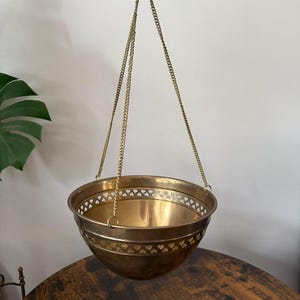 Puede incluir: Un cuenco colgante de latón pulido con un borde decorativo recortado con un patrón de pequeños corazones a lo largo del borde superior. El cuenco está suspendido por tres cadenas finas de latón que se unen en un punto superior central.