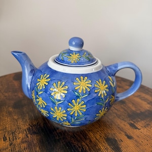 May include: A round ceramic teapot featuring a vibrant blue background with a hand-painted pattern of yellow daisy-like flowers and green stems. The teapot includes a matching lid with a small knob handle, a curved spout, and a side handle.
