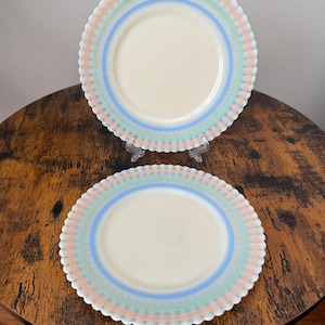 May include: Two off-white plates with scalloped edges and concentric rings of pastel colours: peach, mint green, and blue. The plates are displayed on a dark wooden table, showcasing their decorative design. The plates are a vintage style.