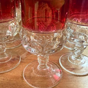 Può includere: Un set di bicchieri da vino vintage con coppe rosso rubino e basi trasparenti decorate. I bicchieri hanno un design classico con piede rotondo e stelo dettagliato. I bicchieri sono disposti su una superficie di legno, mostrando la loro forma elegante.