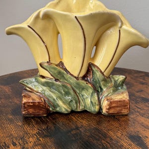 Peut inclure: Un vase en céramique en forme de trois fleurs de lys calla jaune pâle avec des contours bruns. La base présente des feuilles vertes sculptées et des accents rectangulaires bruns. La surface des fleurs jaunes présente un fini craquelé.