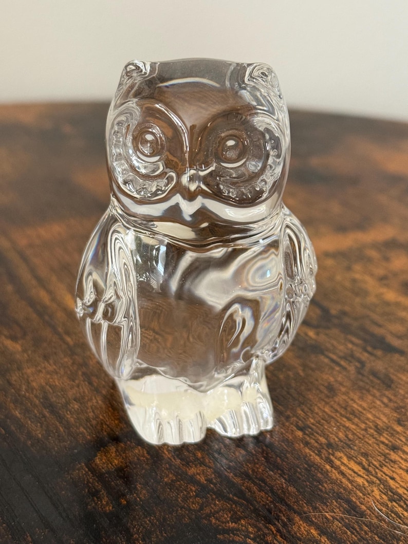 May include: A clear glass figurine shaped like an owl. The piece features detailed molded textures representing feathers on the wings and chest, with prominent circular eyes and small ear tufts at the top of the head, creating a reflective surface.