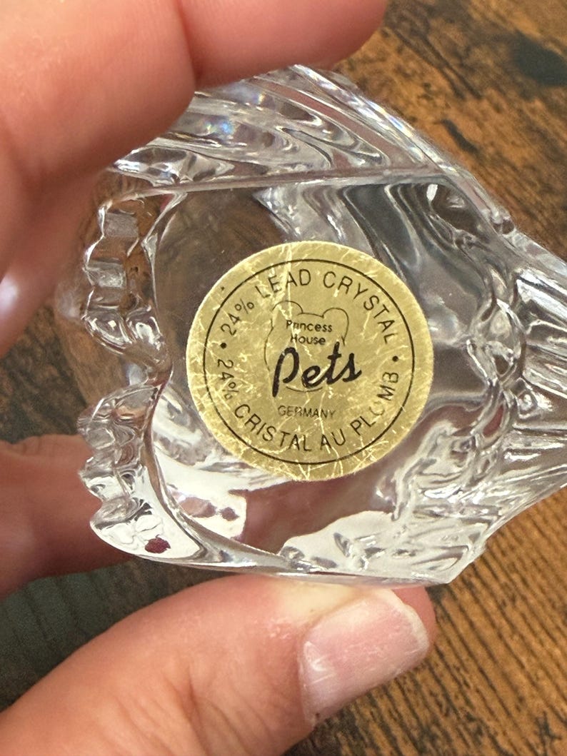 May include: A clear 24% lead crystal figurine with a circular gold sticker on the base. The sticker features the text Princess House Pets, 24% Lead Crystal, 24% Cristal Au Plomb, and Germany. The crystal has a faceted, textured surface design.