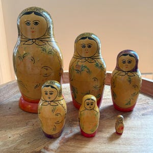 Könnte beinhalten: Ein Set aus sechs Holz-Matrjoschka-Puppen in verschiedenen Größen. Die Puppen sind gelb bemalt und weisen Gesichter, florale Akzente sowie rote und violette Details auf. Die größte Puppe ist etwa 18 cm hoch.