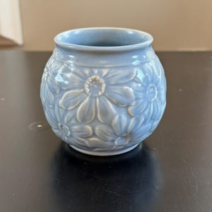 Peut inclure: Petit vase rond en céramique bleu clair avec un motif floral. Le vase présente des fleurs en relief en forme de marguerites et une large ouverture. Il est posé sur une surface sombre.
