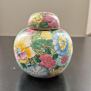 Pode incluir: Um vaso redondo de cerâmica com tampa, adornado com um padrão floral vibrante. O design apresenta flores e folhas rosa, amarelas, azuis e verdes. O vaso é provavelmente usado para fins decorativos.