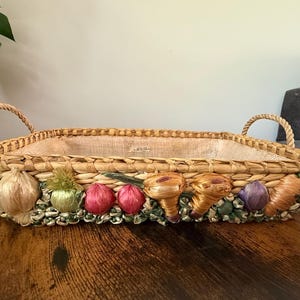 May include: A rectangular woven basket with two rope handles and a light tan fabric liner. The exterior is decorated with a row of colorful, textured vegetable-shaped accents including garlic, onions, and carrots in shades of green, red, and orange.