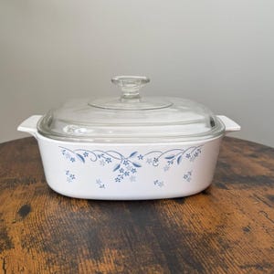 May include: White casserole dish with a clear glass lid. The dish is decorated with a blue floral pattern around the rim. The dish has handles on each side and is sitting on a wooden surface.