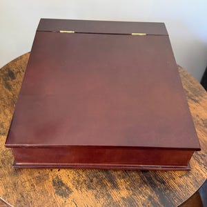 Peut inclure: Une boîte rectangulaire en bois avec une finition acajou foncé et une surface supérieure lisse et plate. La boîte présente deux petites charnières métalliques de couleur dorée visibles le long du bord arrière, reliant le couvercle à la base.