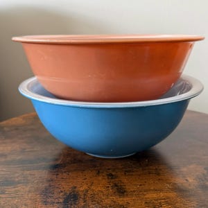 Puede incluir: Dos cuencos mezcladores vintage. El cuenco superior es de color naranja cálido, y el cuenco inferior es azul con un borde blanco. Los cuencos están apilados uno encima del otro y están hechos de un material duradero.
