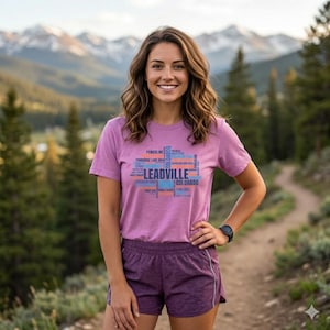 Pode incluir: Uma mulher vestindo uma camiseta e shorts roxos com a palavra "Leadville" em um design retangular. A camisa tem texto adicional em azul, laranja e branco. A mulher está sorrindo e em pé em uma trilha com um cenário de montanha.