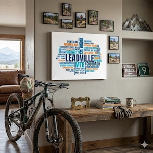 Könnte beinhalten: Eine Innenansicht mit einem Mountainbike, Kunstwerken und dekorativen Elementen. Ein Leinwanddruck zeigt das Wort "Leadville" und andere Texte. Kleine gerahmte Fotos von Mountainbike-Szenen hängen an der Wand. Ein Holztisch enthält Bücher, Handschuhe und eine Tasse.