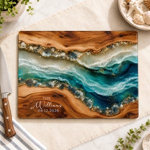 May include: A rectangular wooden cutting board featuring a central resin inlay with swirling shades of teal, blue, and white, accented by gold flecks. The board is engraved with the text "THE Williams 08.12.2026" in a white script and serif font.