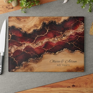May include: A rectangular wooden cutting board featuring a central band of deep red and gold resin with a geode-like texture. The board is personalized with the black text Olivia & Adam 40 Years. A white knife is positioned on the left side.