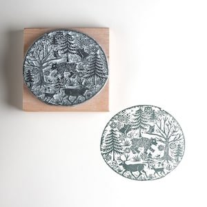 May include: A round, gray stamp with a forest scene featuring a bear, deer, trees, and an owl. The stamp is mounted on a rectangular wooden block. A stamped impression of the design is visible below the stamp.