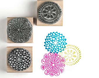 May include: Three wooden blocks with circular rubber stamps featuring intricate floral designs. The stamps are black and gray and are arranged in a row. Below the stamps are three colorful floral designs in shades of blue, pink, and yellow.