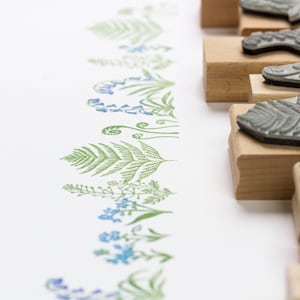 May include: A set of wooden mounted rubber stamps featuring various floral and fern designs. The stamps are arranged in a row on a white surface.