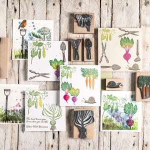 Peut inclure: Une collection de tampons en caoutchouc et leurs impressions à l'encre sur le thème du jardin, incluant des légumes comme des carottes, betteraves et pois, ainsi que des outils, escargots et un oiseau. Une impression porte le texte The land knows you even when you are lost. Robin Wall Kimmerer.