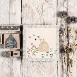 May include: A set of wooden printing blocks with designs of a beehive, bees, flowers, and a border. The blocks are arranged on a white wooden surface.
