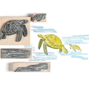 Pode incluir: Carimbos de madeira com desenhos de tartarugas marinhas e padrões de água. Os carimbos mostram uma tartaruga grande, uma tartaruga pequena e padrões de ondas. As imagens carimbadas são em verde, azul e preto.