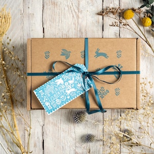 Puede incluir: Una caja de regalo de cartón marrón atada con una cinta y etiqueta color turquesa. La caja tiene ilustraciones turquesas de un pájaro, un conejo y diseños florales. La etiqueta presenta un diseño de arte popular blanco y turquesa de una casa, flores y un conejo.
