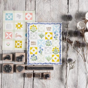 May include: A set of rubber stamps featuring a variety of quilt block designs, including a house, a star, a bunny, a bird, and a flower. The stamps are arranged on a wooden surface with a background of dried flowers and a card with a quilt block design and the text "Love You" and "Well Done".