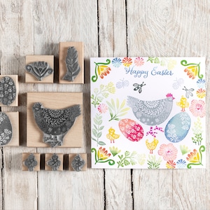 May include: A set of nine wooden stamps featuring Easter-themed designs, including a chicken, eggs, and flowers. The stamps are arranged on a wooden surface with a card featuring a similar design in the background. The card reads "Happy Easter".