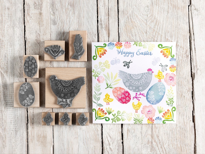 May include: A set of nine wooden stamps featuring Easter-themed designs, including a chicken, eggs, and flowers. The stamps are arranged on a wooden surface with a card featuring a similar design in the background. The card reads "Happy Easter".