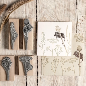 Op de afbeelding: Houten stempels met botanische ontwerpen, waaronder bladeren en zaaddozen, samen met bedrukte kaarten met vergelijkbare illustraties. De stempels zijn gemonteerd op rechthoekige houten blokken. De kaarten en stempels zijn in bruin- en beigetinten.