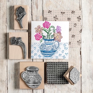 Puede incluir: Una colección de sellos florales y una imagen estampada de un jarrón con flores. Los sellos están montados sobre bloques de madera. La imagen estampada presenta un jarrón azul y blanco con flores rosas y moradas.