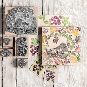 May include: A collection of wooden stamps with nature-themed designs, including a hedgehog, mushrooms, and various leaves and berries. The stamps are arranged with printed cards featuring similar illustrations in shades of green, purple, and yellow. The background is a white wooden surface.