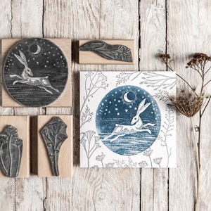 May include: A selection of wooden stamps and a print depicting a running hare under a crescent moon and stars. The stamps feature botanical and hare designs. The print is on a white background with blue and grey accents.