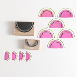 Può includere: Un set di timbri in legno a forma di fette di anguria. I timbri vengono utilizzati per creare fette di anguria rosa con semi neri. I timbri sono disposti su una superficie bianca.