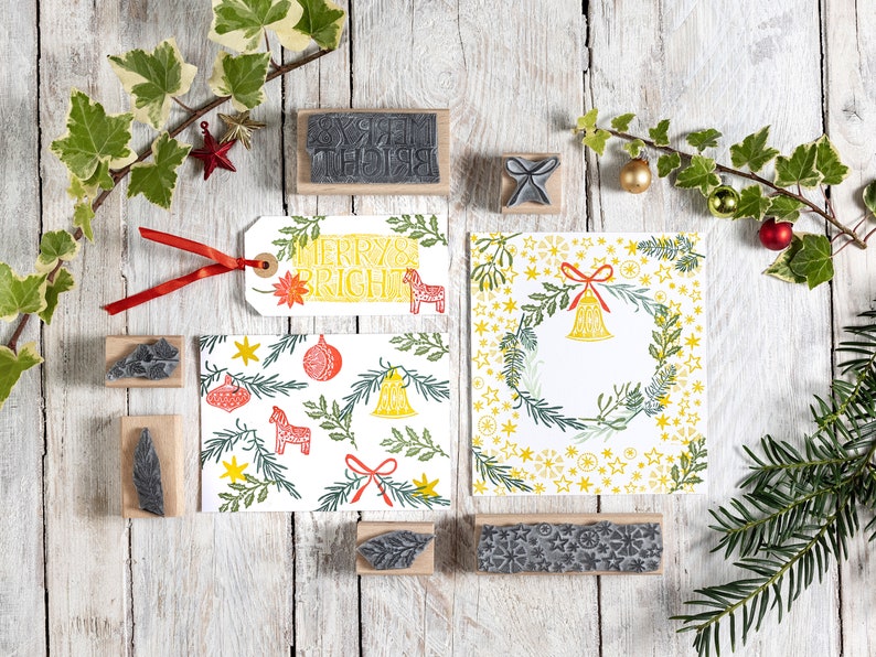 May include: A set of Christmas-themed rubber stamps with designs including a wreath, a bell, a star, a horse, and the words "Merry Bright". The stamps are on a white wooden surface with green ivy and a red ribbon.
