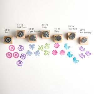 May include: Set of 7 rubber stamps with different designs, including a spot, folk flower, open butterfly, curly, daisy, tiny bird, and side butterfly. The stamps are arranged in a row on a white surface, with the corresponding stamped images below each stamp.