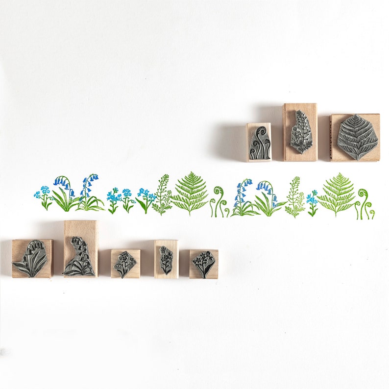 May include: A set of 12 wooden stamps featuring various floral and fern designs. The stamps are arranged in a row on a white background. The designs include bluebells, ferns, and leaves.