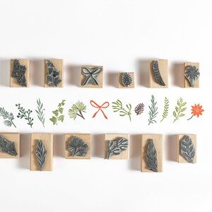 May include: A set of 16 wooden stamps featuring various botanical designs, including leaves, branches, and a red bow. The stamps are arranged in two rows on a white background.