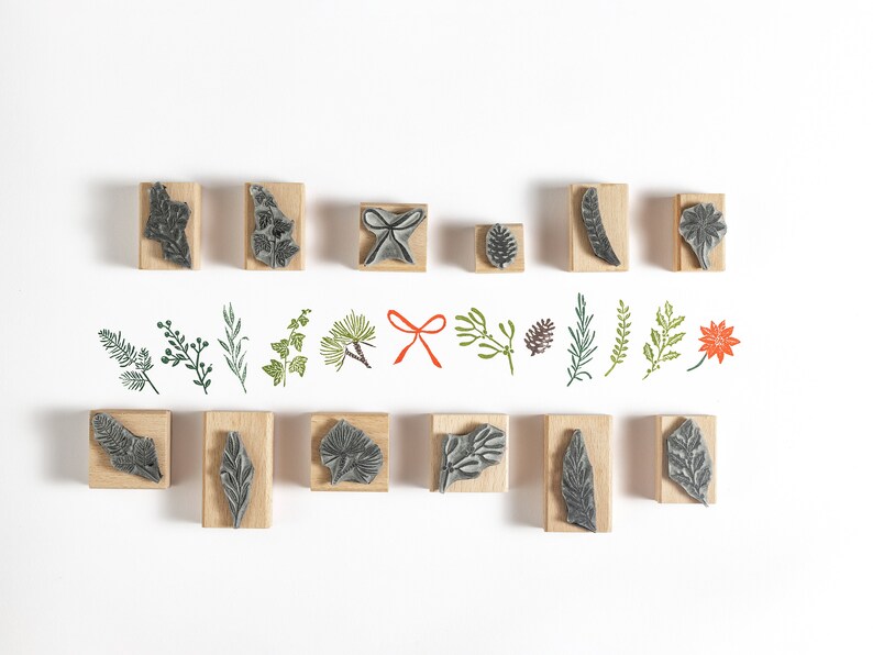 May include: A set of 16 wooden stamps featuring various botanical designs, including leaves, branches, and a red bow. The stamps are arranged in two rows on a white background.
