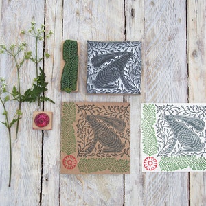 Puede incluir: Un surtido de materiales de arte hechos a mano, que incluyen un sello de conejo tallado, un sello de helecho y tres impresiones cuadradas con un diseño de conejo. Las impresiones son en tonos de gris, marrón y verde, con un borde floral.