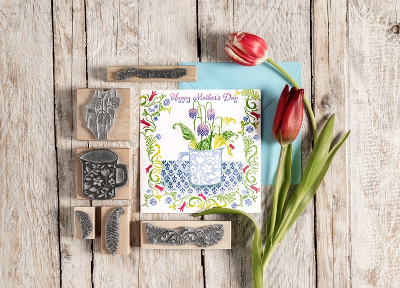 May include: A set of wooden stamps with floral and leaf designs, including a mug with flowers, a fern, and a leaf. The stamps are arranged on a wooden surface with a card that reads "Happy Mother's Day" and a bouquet of red tulips.