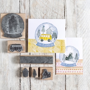 Op de afbeelding: Een set houten gemonteerde rubberen stempels met een winterthema. De stempels omvatten een sneeuwbol met een gele bestelbus erin, een sneeuwbol met een huis erin, een bestelbus, een huis, twee bomen en een gepatenteerde rand. De stempels bevinden zich op een witte houten achtergrond.