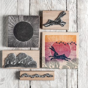 May include: A selection of woodblock prints and stamps with nature themes. The prints depict a sunburst, a running hare, trees, and a landscape with birds. The stamps are mounted on wooden blocks, presented on a wooden surface.