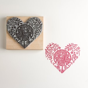 Può includere: Un timbro in gomma a forma di cuore con un disegno in bianco e nero di un albero con uccelli e pere. Il timbro è montato su un blocco di legno.
