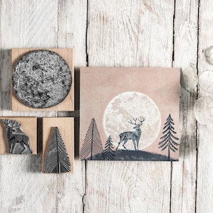 May include: A set of four wooden stamps featuring a deer, a tree, and a moon. The stamps are on a white wooden background.