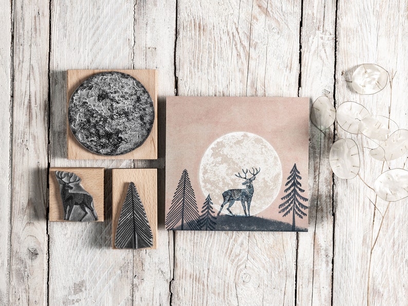 May include: A set of four wooden stamps featuring a deer, a tree, and a moon. The stamps are on a white wooden background.